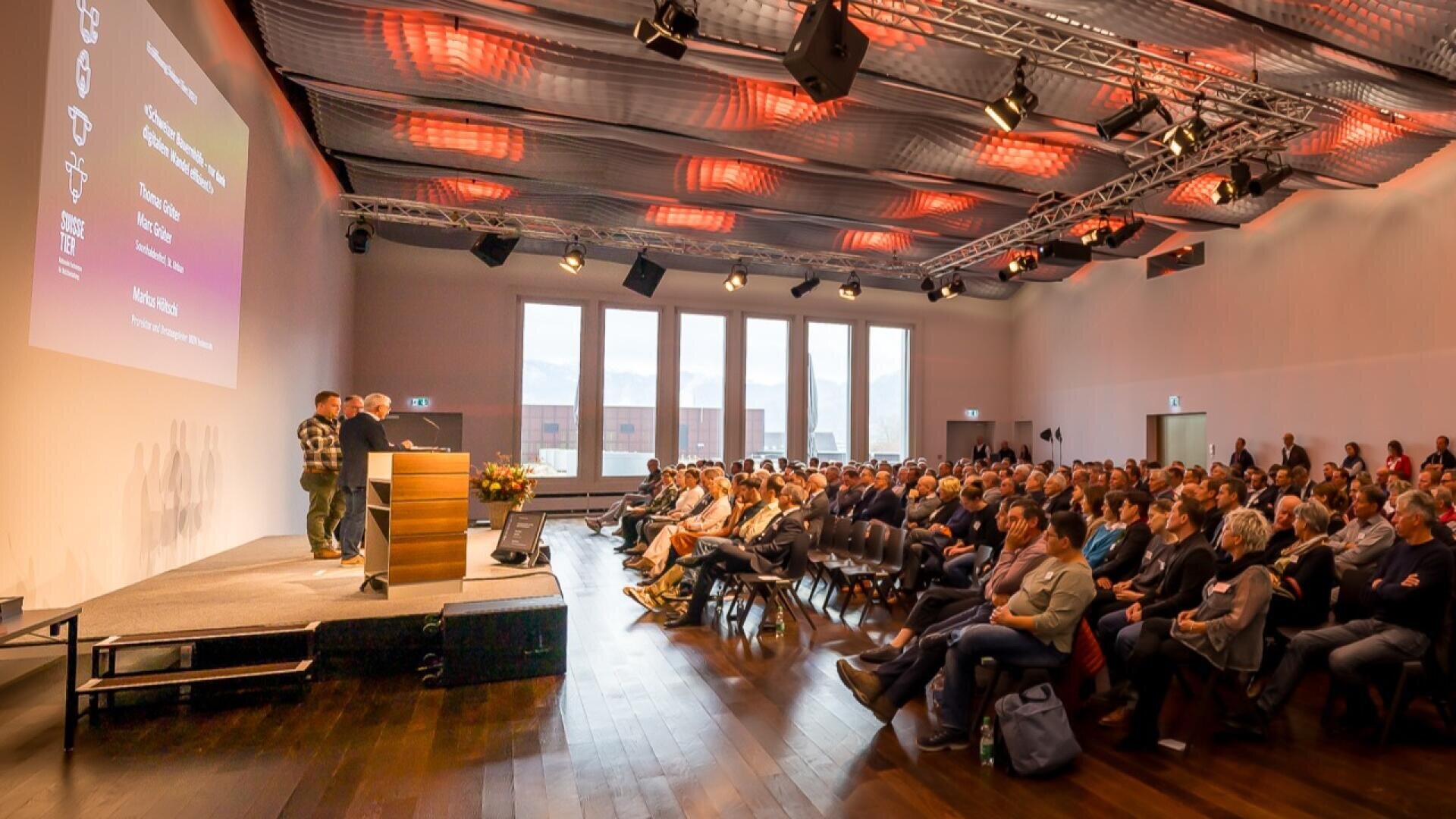 Eine große Gruppe von Personen sitzt während einer Präsentation in einem modernen Konferenzraum mit Holzboden und gemusterter Deckenbeleuchtung in Reihen vor einer Bühne. Einige Teilnehmer stehen an den Seiten- und Rückwänden.