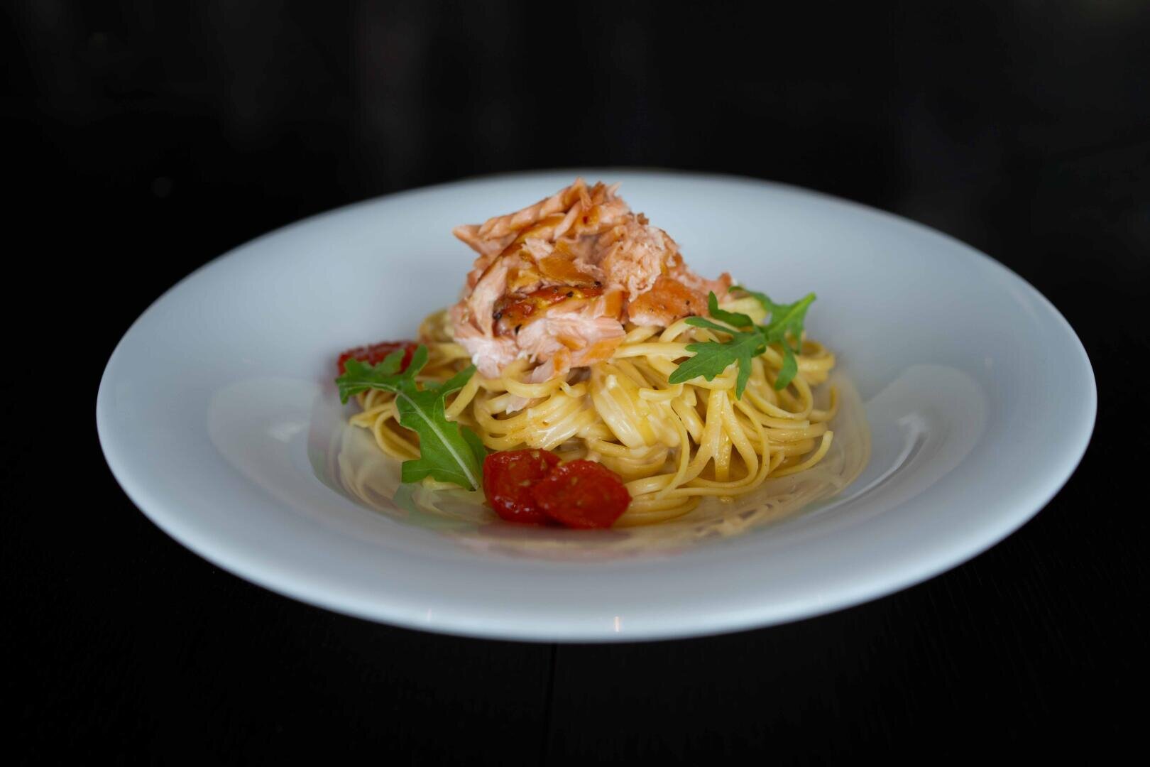 Ein weißer Teller mit Spaghetti, die mit Lachsflocken, Kirschtomaten und Rucola belegt sind, vor einem dunklen, unscharfen Hintergrund.