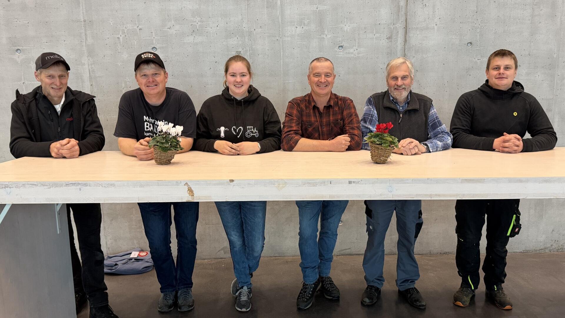 Sechs Personen sitzen nebeneinander hinter einem hellen Tisch mit zwei kleinen Topfpflanzen und lächeln in die Kamera. Im Hintergrund ist eine schlichte graue Betonwand zu sehen.