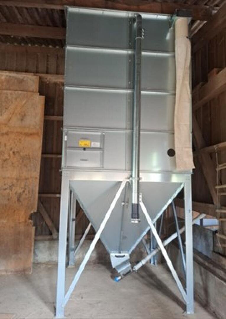 Ein großes Futtersilo aus Metall mit trichterförmigem Boden, das auf vier Beinen in einer Holzscheune steht. An der Seite des Silos verläuft ein senkrechtes Rohr, und oben rechts ist ein flexibler Schlauch angebracht.