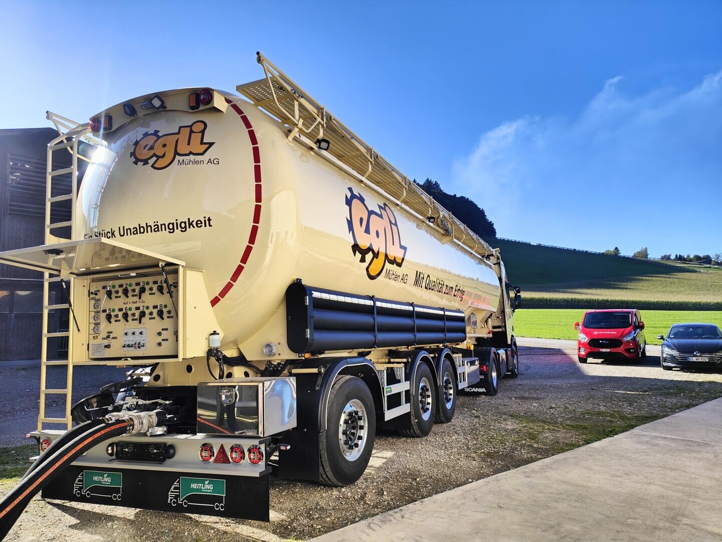 Ein großer gelber Egli-Tankwagen steht auf einer gepflasterten Fläche in der Nähe eines grasbewachsenen Feldes und eines blauen Himmels, mit einem roten Lieferwagen und Autos im Hintergrund. An der Rückseite des Lkw sind Schläuche angeschlossen.