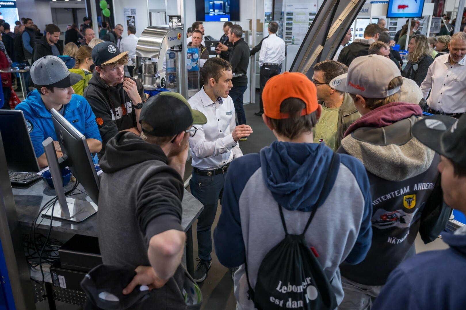 Eine Gruppe junger Leute versammelt sich um einen Mann, der ein technisches Gerät auf einer belebten Ausstellung oder Messe erklärt. Im Hintergrund sind verschiedene Displays, Bildschirme und andere Teilnehmer zu sehen.