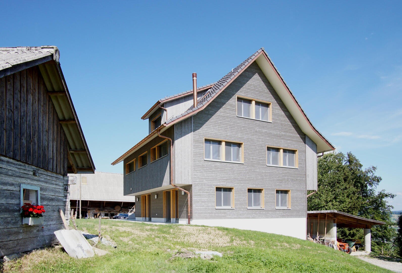 Ein modernes dreistöckiges Haus mit grauen Holzverkleidungen und einem steilen roten Ziegeldach steht auf einem grasbewachsenen Hang neben einer hölzernen Scheune unter einem klaren blauen Himmel.
