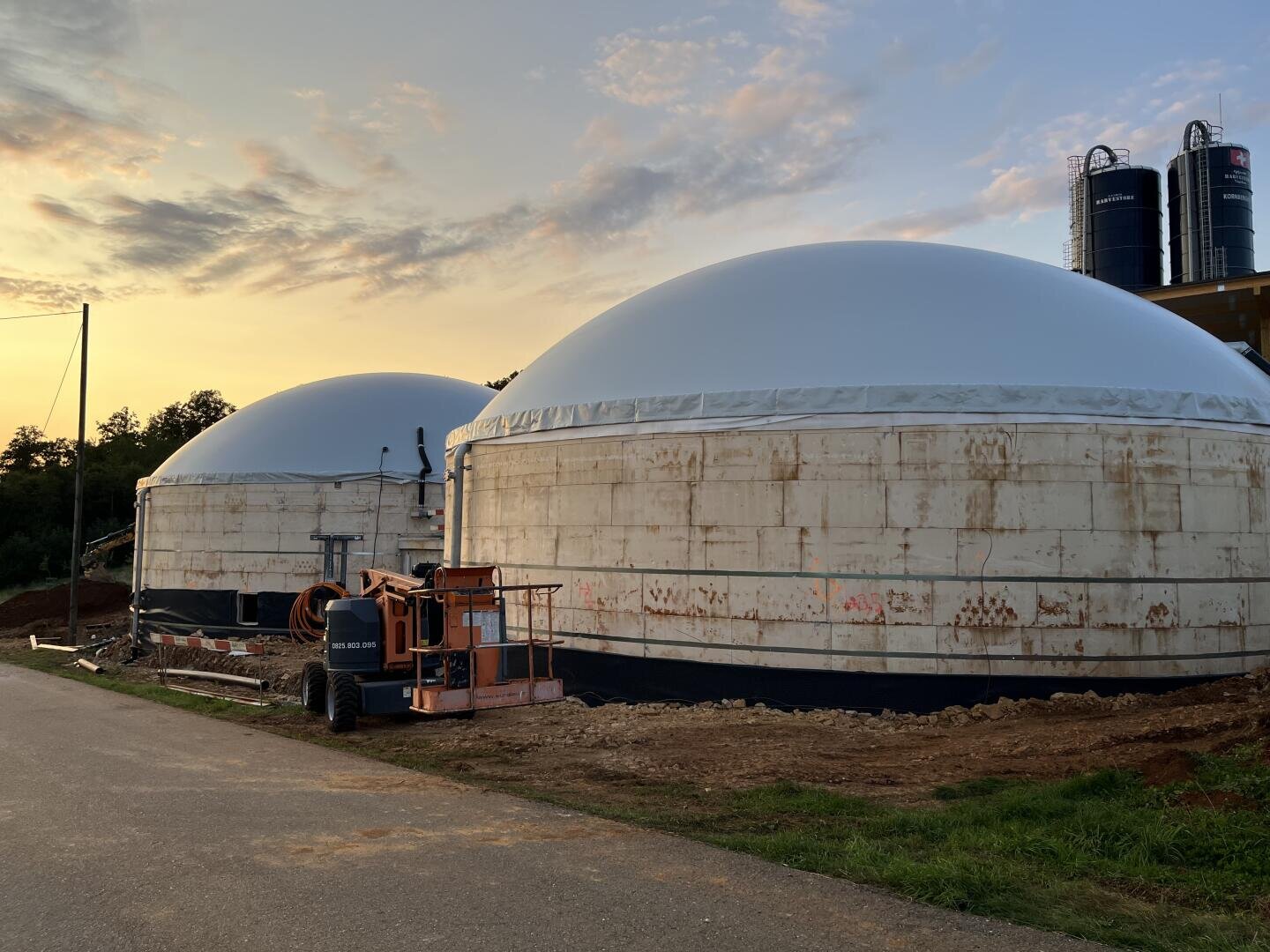 Zwei große, weiße Industrietanks stehen bei Sonnenuntergang auf einem unbefestigten Grundstück in der Nähe einer Straße, daneben ist ein orangefarbener Aufzug geparkt, und im Hintergrund sind bei teilweise bewölktem Himmel weitere Silos zu sehen.