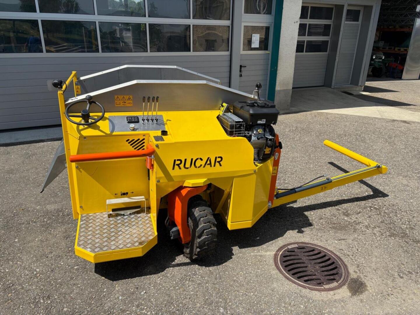Ein gelbes Industriefahrzeug mit der Aufschrift RUCAR, das mit einem Lenkrad, einem Motor und einer Metallstufe ausgestattet ist, steht an einem sonnigen Tag vor einem Gebäude mit Garagentoren.
