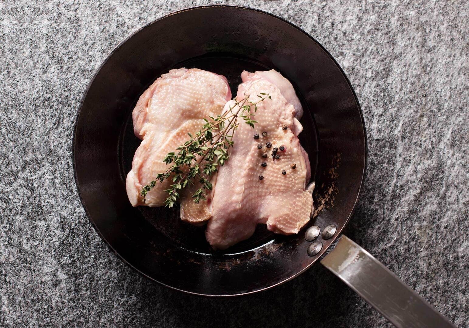Rohe Hühnerbrüste mit Thymianzweigen und ganzen Pfefferkörnern in einer schwarzen Bratpfanne auf einer grauen Steinfläche anrichten.