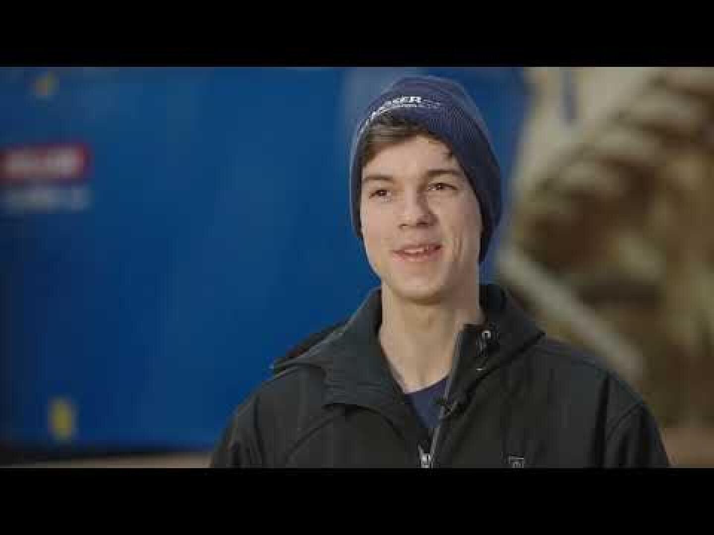 Eine junge Person, die eine blaue Mütze und eine schwarze Jacke trägt, lächelt, während sie im Freien steht, mit einem blauen Hintergrund und einigen unscharfen Objekten im Hintergrund.