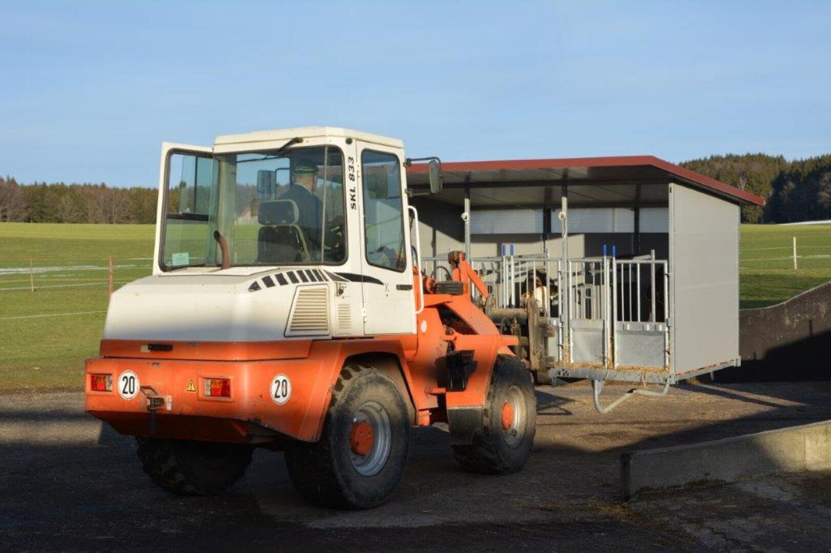 Ein orange-weißer Frontlader hebt eine große Metallkonstruktion mit rotem Dach auf einer Landstraße an, mit grünen Feldern und Bäumen im Hintergrund.