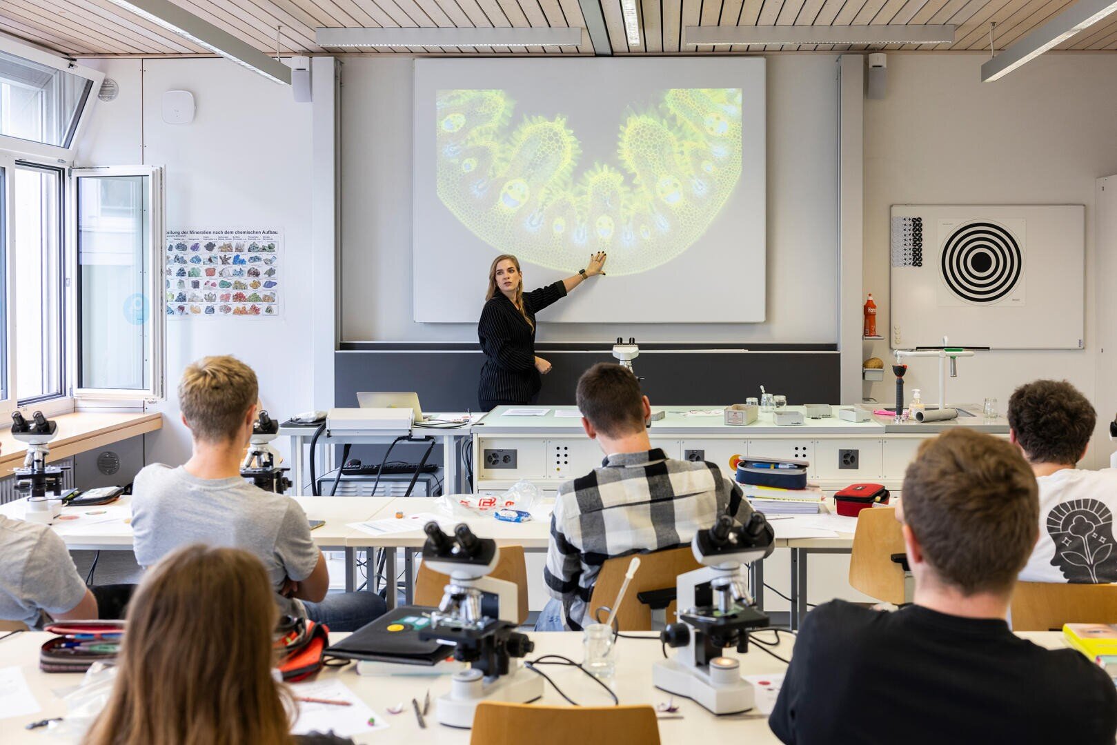 Ein Lehrer zeigt auf eine Nahaufnahme des Querschnitts eines Pflanzenstamms, die auf eine Leinwand projiziert wird, während die Schüler in einem Klassenzimmer mit Mikroskopen zuhören und Notizen machen.