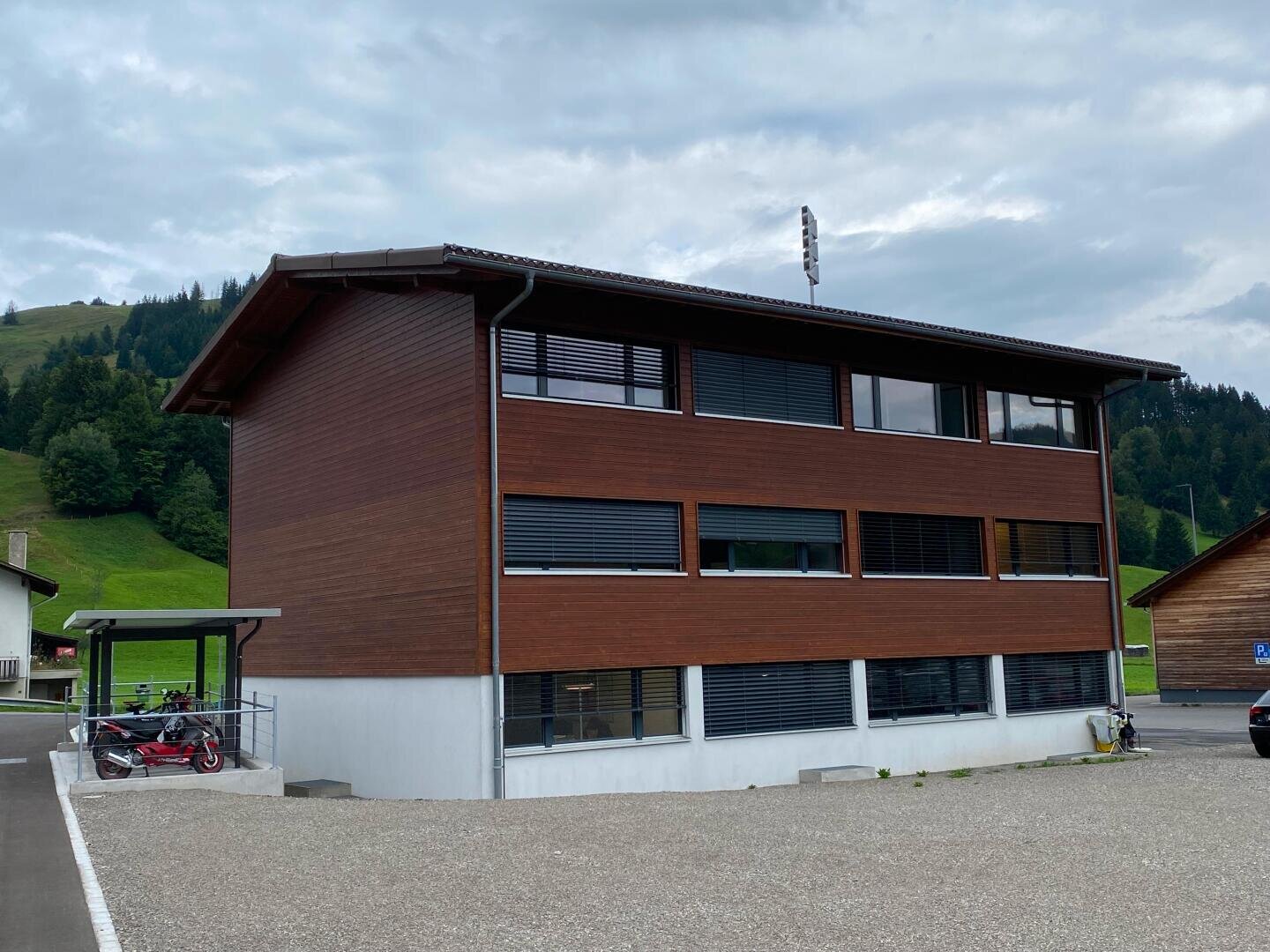 Ein modernes, dreistöckiges Gebäude mit Holzverkleidung und großen Fenstern steht neben einer Kiesfläche, mit grünen Hügeln und Bäumen im Hintergrund unter einem bewölkten Himmel. Unter einem kleinen Unterstand in der Nähe sind Fahrräder abgestellt.