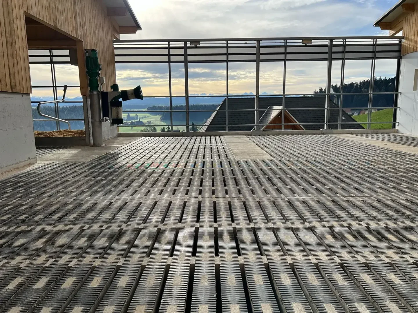 Metallspaltenboden im Inneren einer großen Scheune mit offenen Seiten, mit Blick auf eine malerische ländliche Landschaft mit Hügeln, Feldern und Bäumen unter einem teilweise bewölkten Himmel.