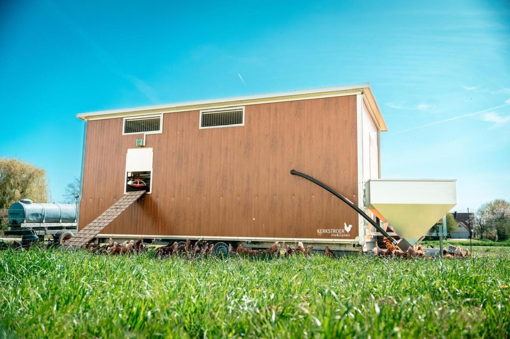 Ein großer, moderner Hühnerstall aus Holz auf Rädern steht auf grünem Gras unter einem strahlend blauen Himmel, mit Hühnern, die draußen herumlaufen, und einem weißen Futterbehälter in der Nähe.