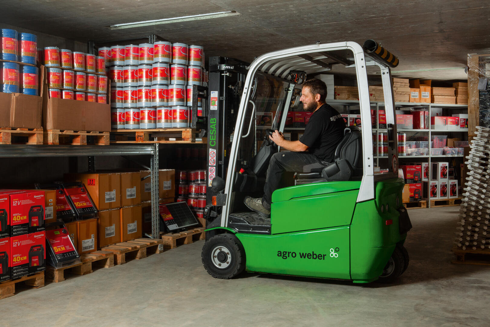 Ein Mann bedient einen grünen Gabelstapler mit der Aufschrift agro weber in einem Lagerhaus und bewegt Gegenstände in der Nähe von Regalen, die mit Farbdosen, Kisten und verschiedenen Produkten bestückt sind.