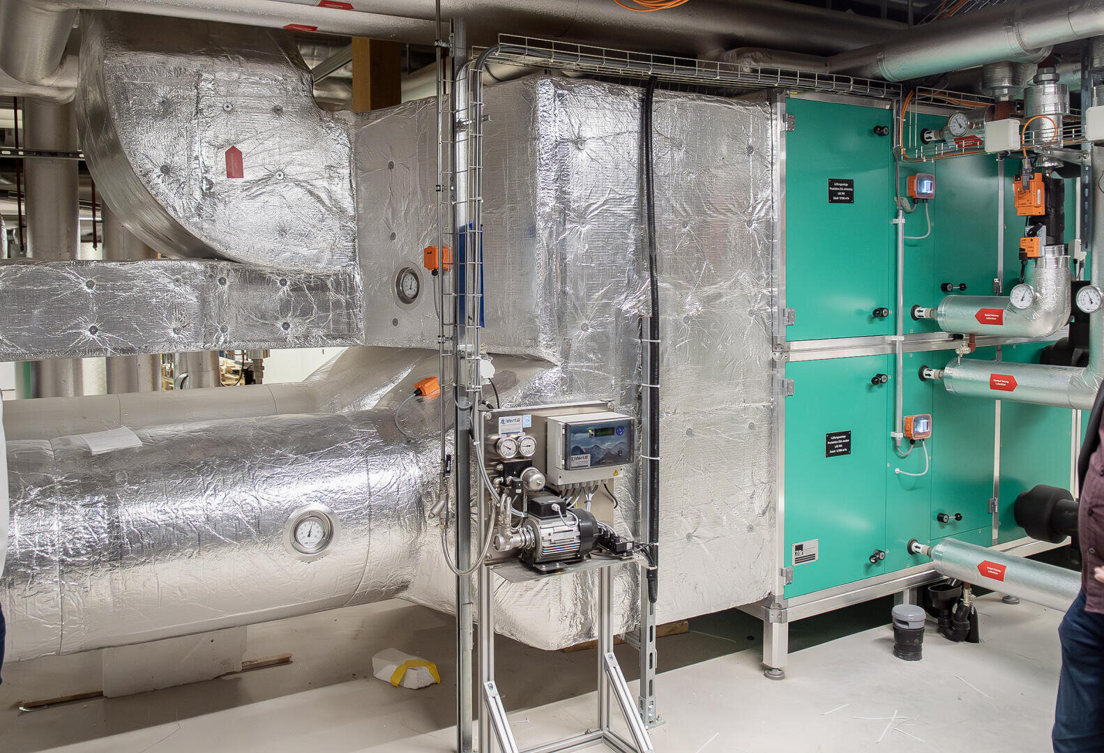 Großes industrielles HLK-System mit isolierten Metallkanälen und einem grünen Lüftungsgerät, umgeben von Rohren, Messgeräten und Schalttafeln in einem Maschinenraum.