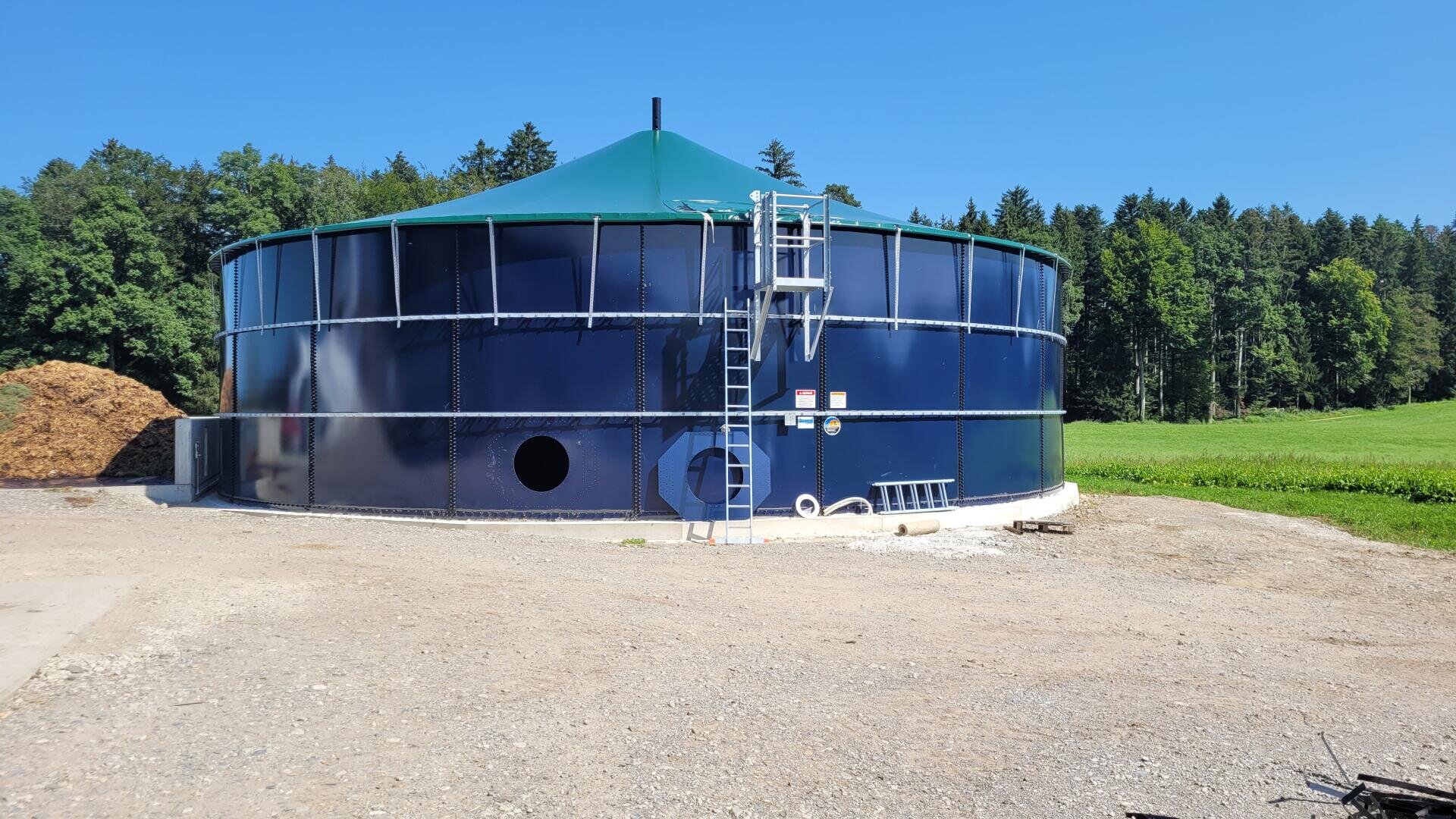 Ein großer, runder, blauer Metalltank mit einem grünen Kegeldach steht im Freien auf Kies, umgeben von grünem Gras und Bäumen. An der Seite sind eine Metallleiter und ein Sicherheitsgeländer angebracht. Der Himmel ist klar und blau.