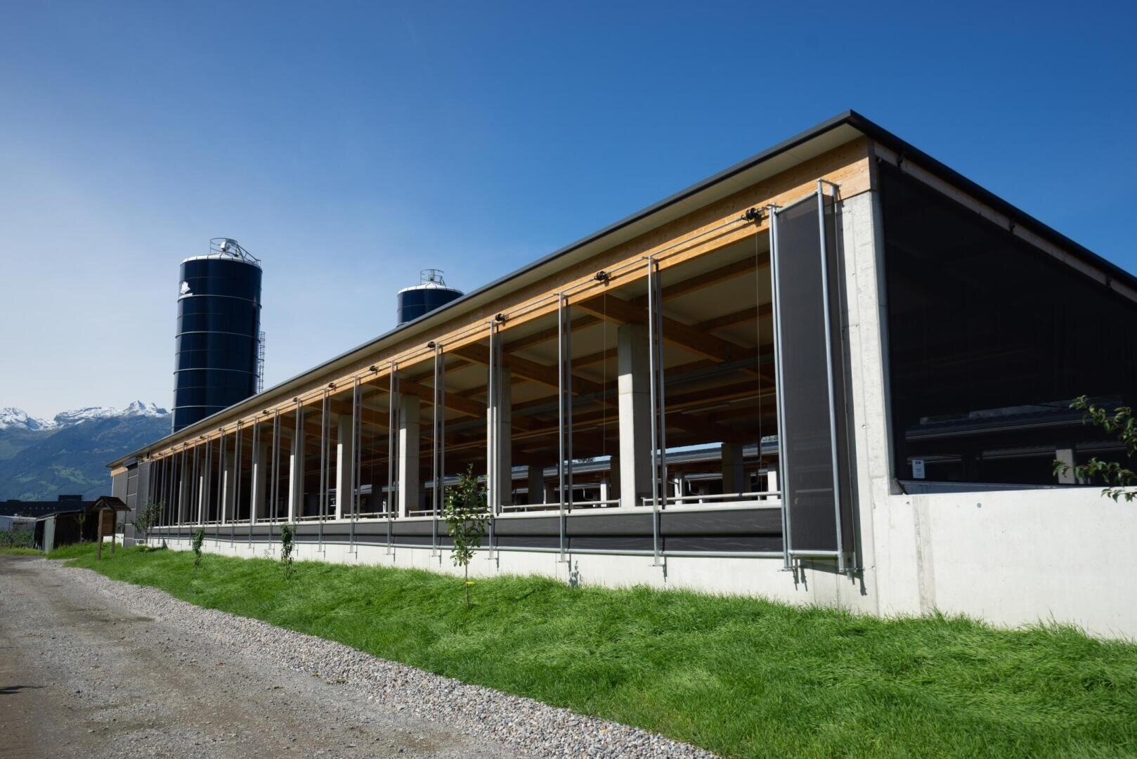 Ein modernes landwirtschaftliches Gebäude mit offenen Seiten, einem Holzdach und großen schwarzen Silos im Hintergrund, auf einem Schotterweg mit grünem Gras und Bergen in der Ferne unter einem klaren blauen Himmel.