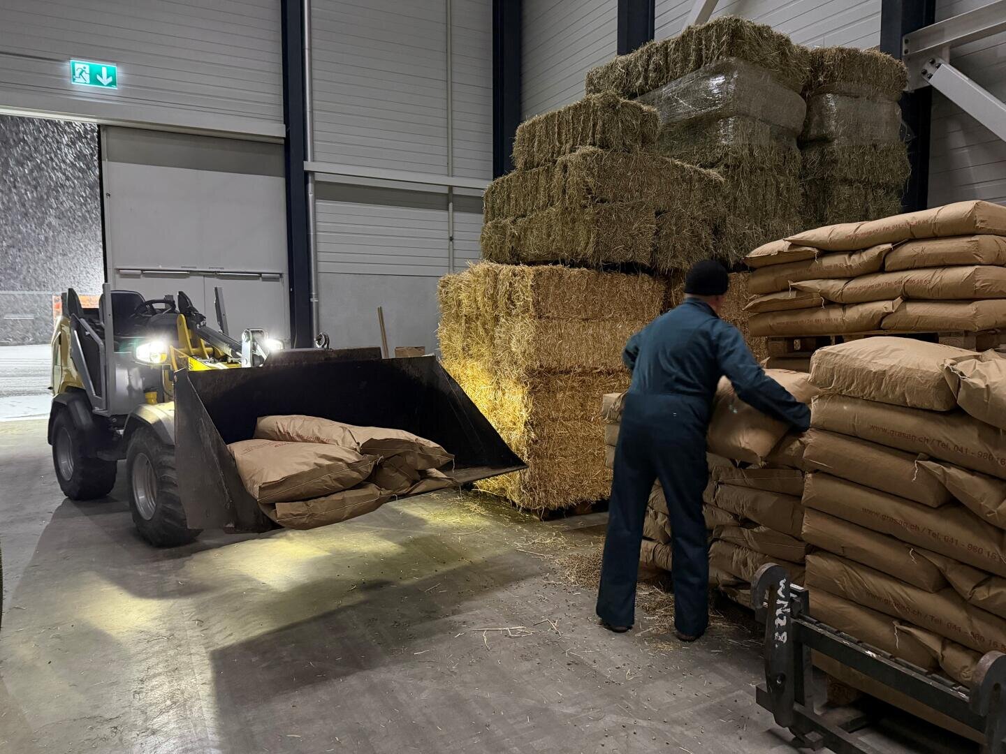 Ein Mann in einem Overall stapelt große Papiersäcke neben Heuballen in einem Lagerhaus, während ein kleiner Lader weitere Säcke von draußen durch eine offene Tür liefert.