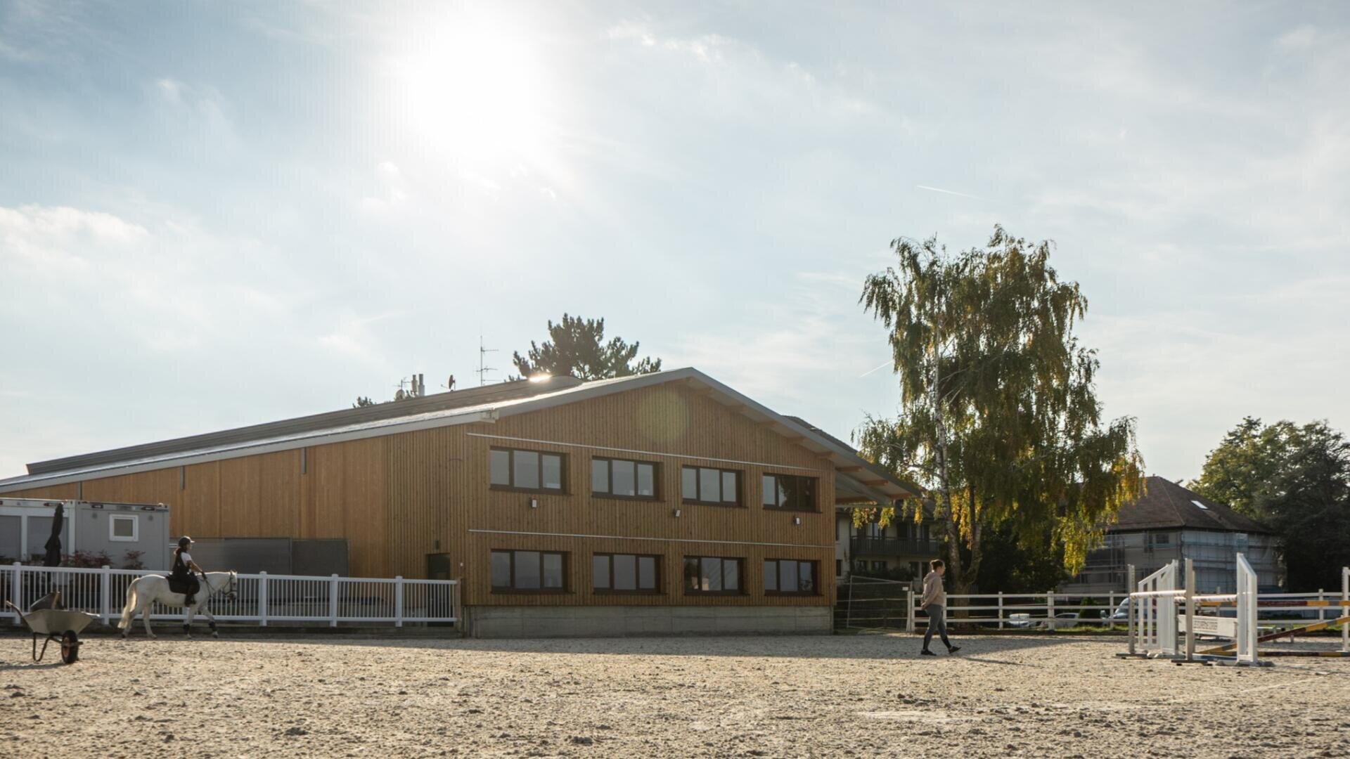 Ein sonniger Reitplatz im Freien mit einem modernen Holzgebäude im Hintergrund. Eine Person reitet ein weißes Pferd auf der linken Seite, und eine andere Person führt ein braunes Pferd auf der rechten Seite. Weiße Zäune und Bäume umgeben das Gelände.