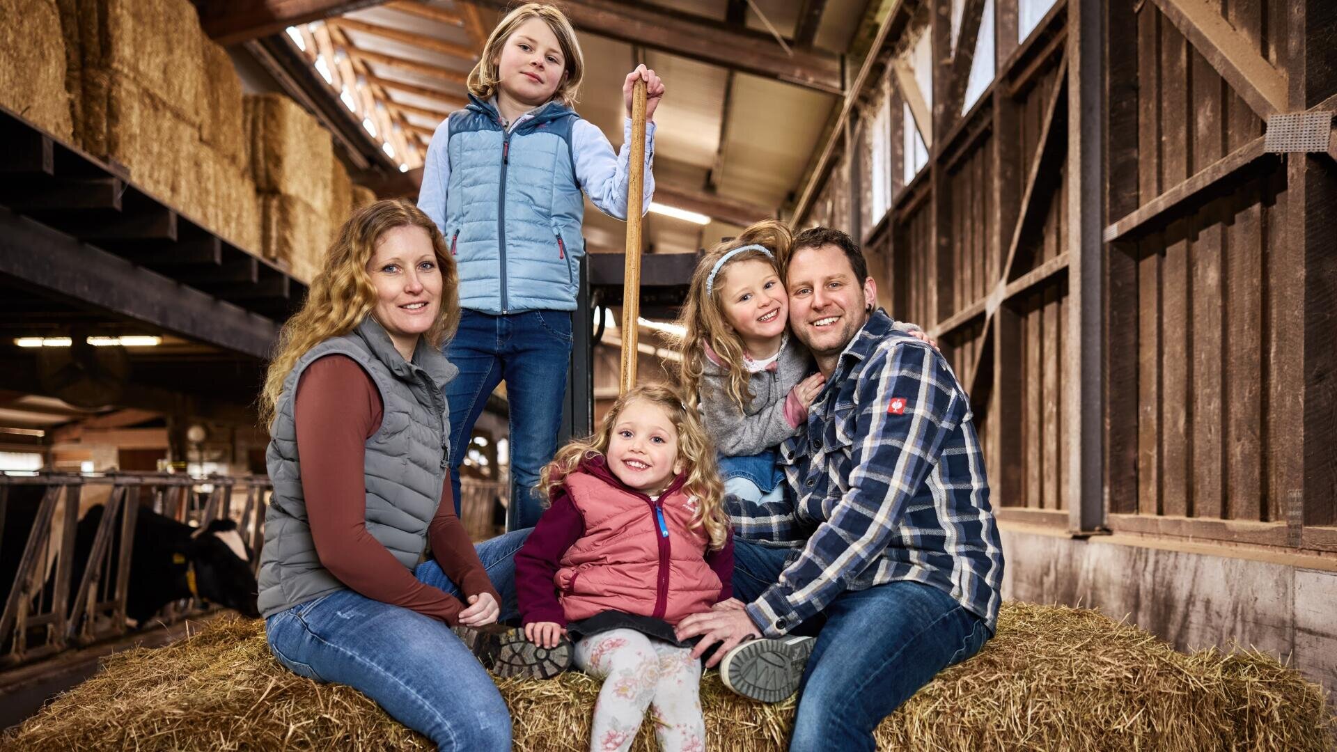 Eine fünfköpfige Familie in legerer Winterkleidung sitzt auf Heuballen in einer Scheune und lächelt gemeinsam. Das von oben einfallende Sonnenlicht hebt die rustikale Holzeinrichtung und das gestapelte Heu im Hintergrund hervor.