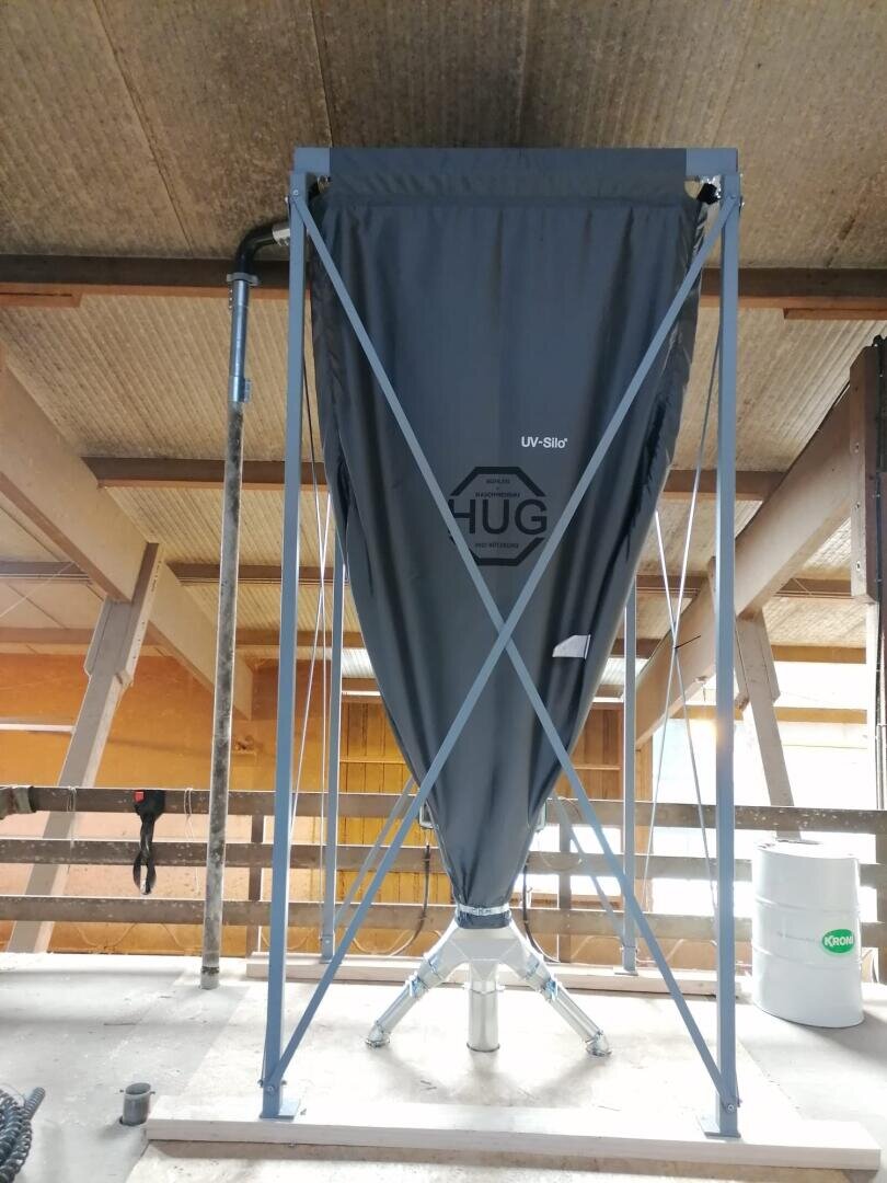 Ein großes, graues Stoffsilo mit der Aufschrift HUG steht in einem Metallrahmen innerhalb einer Scheune oder eines Industriegebäudes; im Hintergrund sind Rohre und Holzbalken zu sehen.