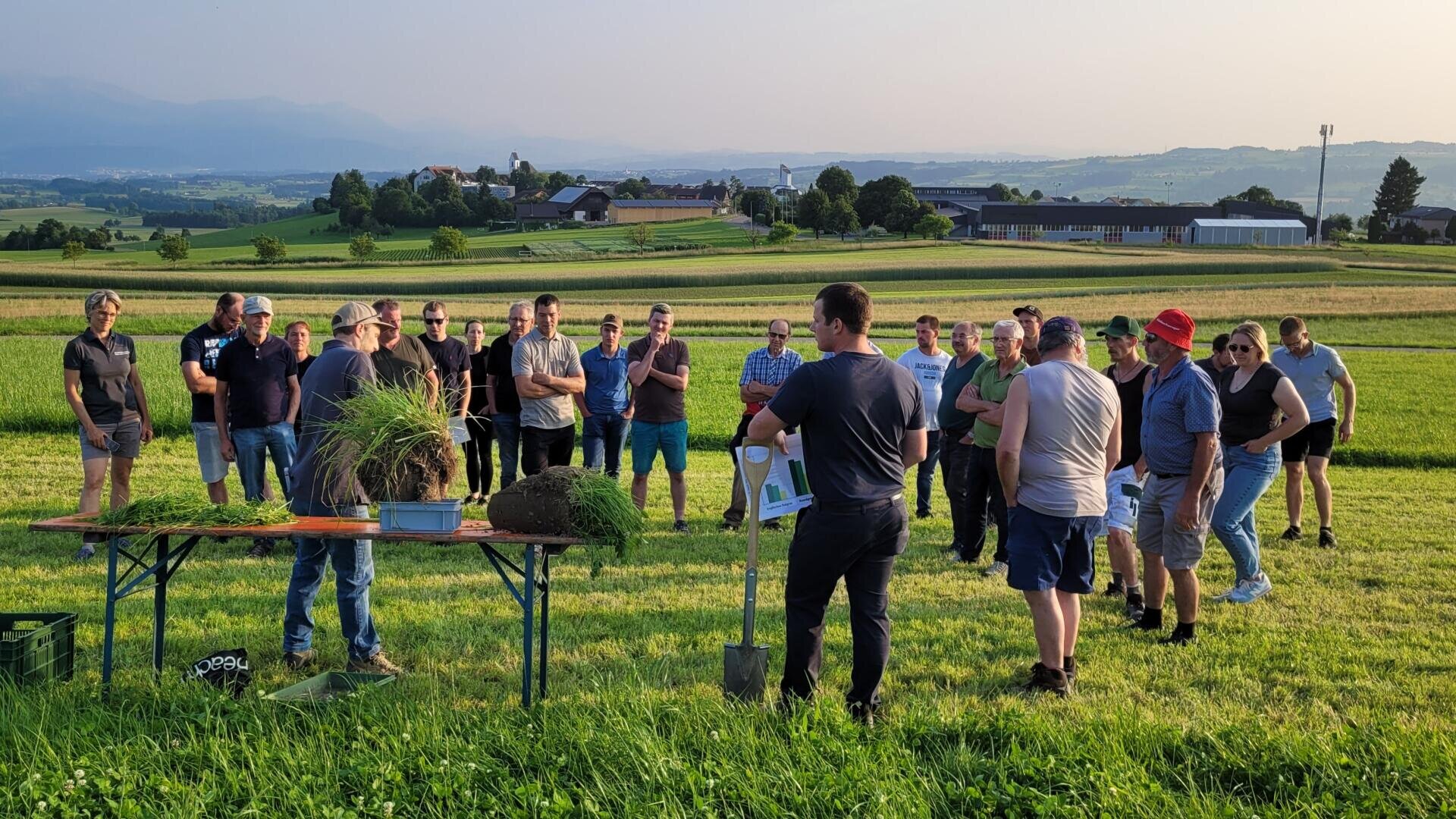 Eine Gruppe von Menschen steht auf einer Wiese und hört aufmerksam einem Mann zu, der eine Demonstration mit Erde und Gras auf einem Tisch gibt. Im Hintergrund sind bei klarem Himmel ländliche Gebäude und sanfte Hügel zu sehen.