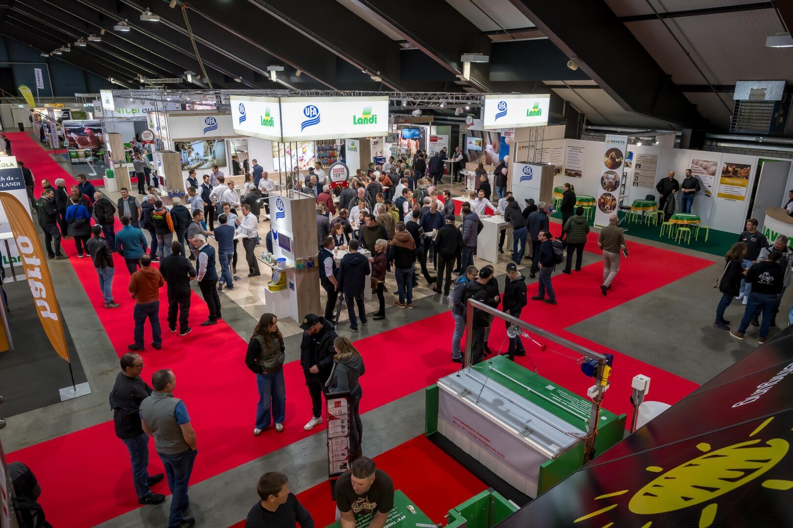 Eine belebte überdachte Messe mit vielen Menschen, die auf einem roten Teppich um die verschiedenen Messestände herumgehen und sich versammeln. Die Halle ist gut beleuchtet und die Firmenschilder und Produktdisplays sind überall sichtbar.