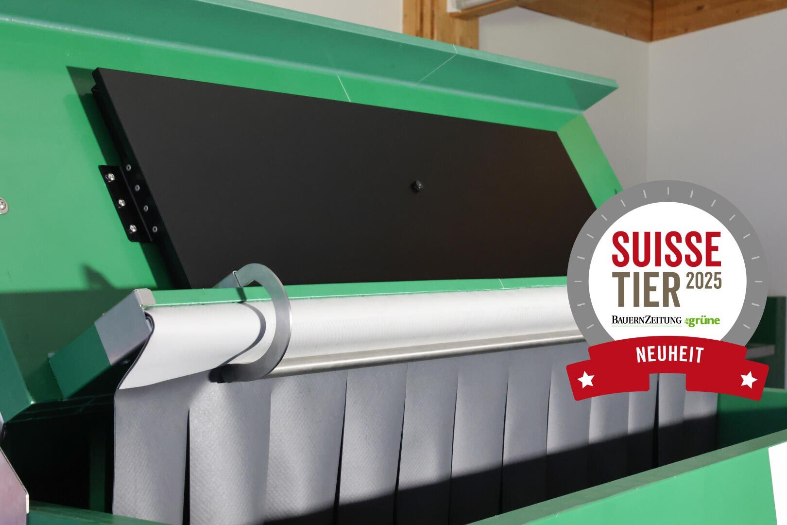 Nahaufnahme einer grünen Industriemaschine mit einer schwarzen Tafel und einem Sprossenförderer im Inneren. Auf der rechten Seite befindet sich ein kreisförmiges Schild mit der Aufschrift Suisse Tier 2025 Neuheit und den Logos von BauernZeitung und Grüne.