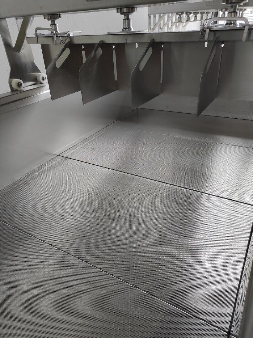 Nahaufnahme einer industriellen Maschine aus rostfreiem Stahl mit Metallschaufeln und einem perforierten Förderband oder einer perforierten Oberfläche, die wahrscheinlich in der Lebensmittelverarbeitung oder -herstellung eingesetzt wird.