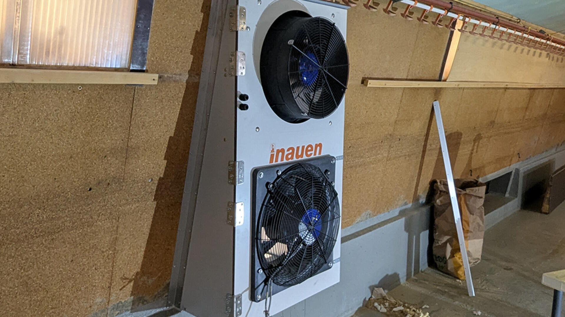 In einem Gebäude mit braunen Korkwänden und freiliegenden Rohren ist ein weißes, an der Wand montiertes Kühlgerät mit zwei großen schwarzen Lüftern mit der Aufschrift "inauen" installiert. Auf dem Boden in der Nähe sind Trümmer und eine Papiertüte zu sehen.