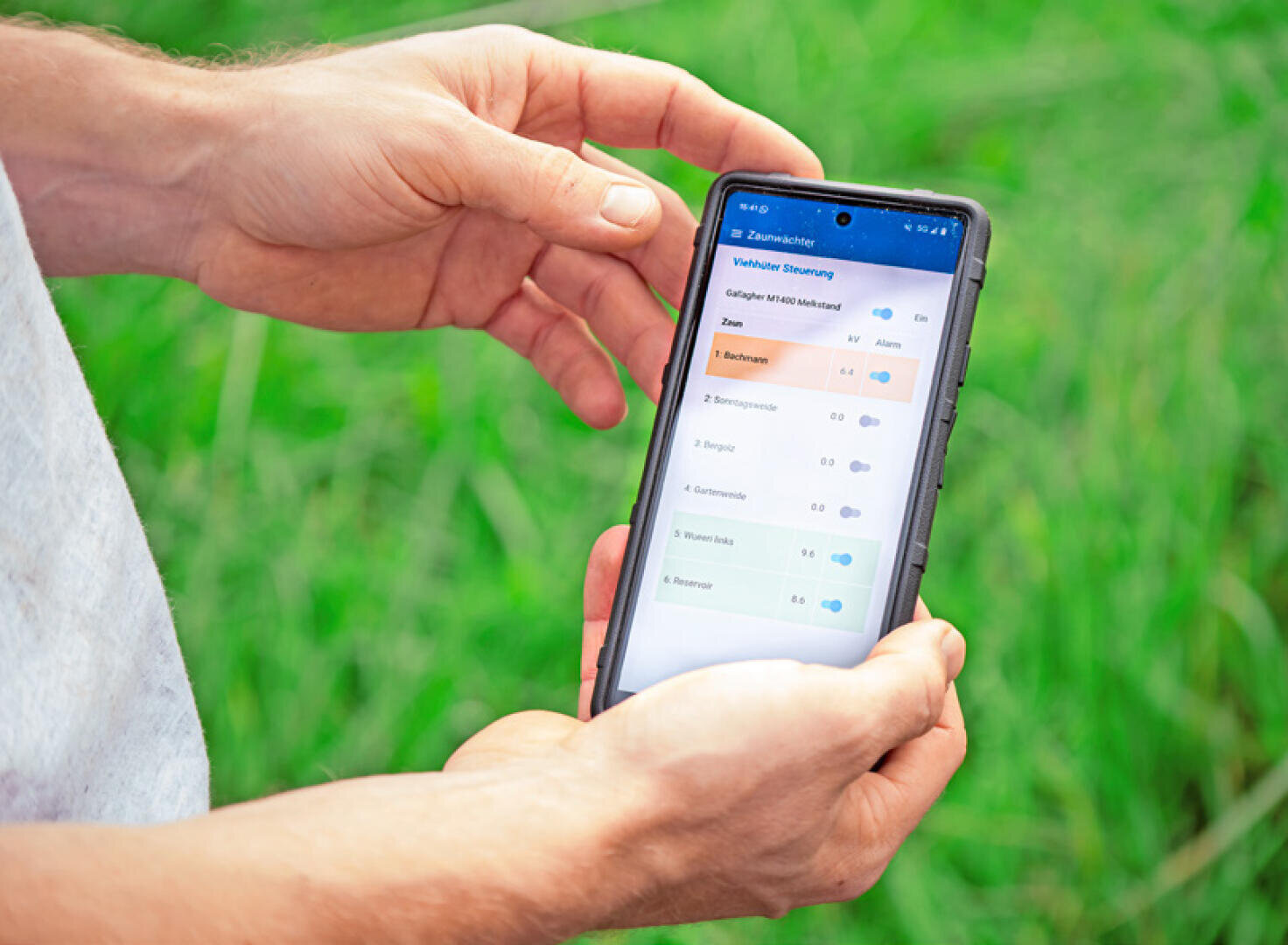 Eine Person hält ein Smartphone im Freien vor einem grasbewachsenen Hintergrund und zeigt eine App mit verschiedenen anpassbaren Einstellungen und farbigen Schiebereglern an.