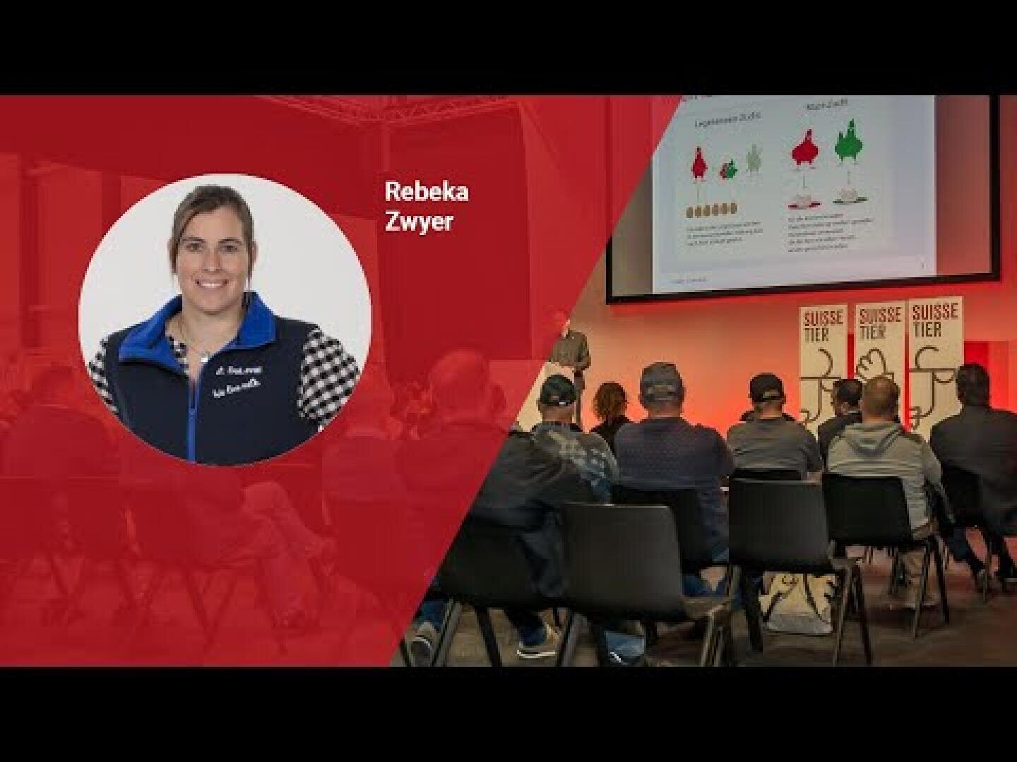 Es findet eine Präsentation statt, bei der das Publikum vor einer Bühne sitzt, auf der Diagramme auf eine Leinwand projiziert werden. Ein Einschub zeigt eine Frau namens Rebeka Zwyer, die lächelt und eine blau-schwarze Jacke trägt.