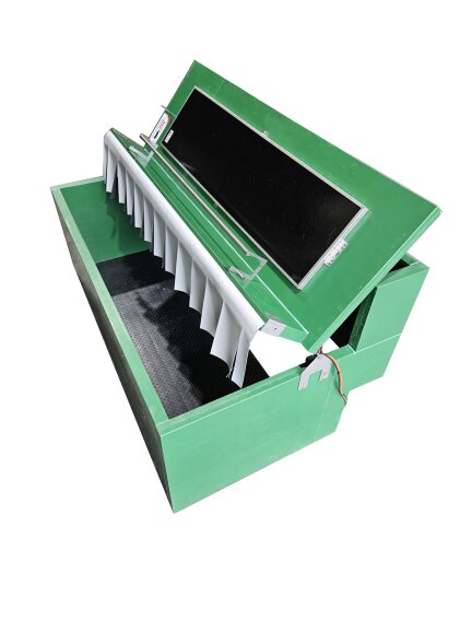 Ein grüner, rechteckiger Kasten mit einem geöffneten Scharnierdeckel, der ein schwarzes Fach im Inneren freigibt. An dem Deckel hängen weiße vertikale Trennwände oder Klappen, und an der Unterseite des Deckels ist ein elektronisches Bauteil angebracht.