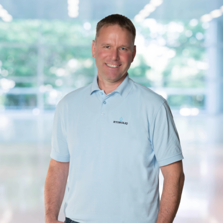 Ein Mann in einem hellblauen Poloshirt mit dem Aufdruck STENGLAGD steht in einem Haus und lächelt in die Kamera. Der Hintergrund ist leicht unscharf und zeigt große Fenster mit Grünpflanzen draußen.