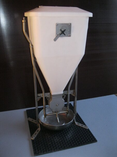 Ein weißer, trichterförmiger Kunststoffspender mit Metallhebel und -rahmen, der auf einer schwarzen Matte steht. Unten befindet sich ein Ausguss, der zu einer runden Metallschale führt.