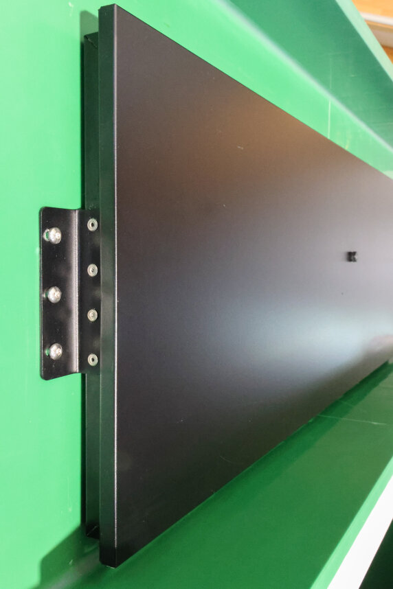 Ein schwarzes Metallpaneel mit einer durch Schrauben befestigten Halterung ist auf einer grünen Fläche angebracht, die aus einem Winkel betrachtet wird, der die Länge des Paneels und seine glatte, matte Oberfläche hervorhebt.