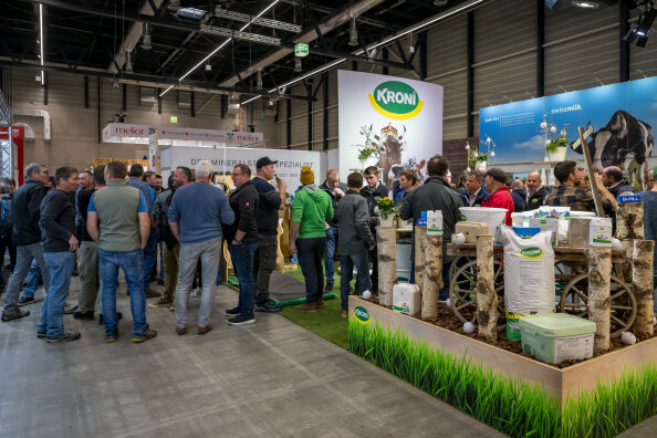 Eine Menschenmenge versammelt sich um den KRONI-Stand auf einer überdachten Landwirtschaftsmesse. Der Stand zeigt Bilder von Kühen, grünes Branding, landwirtschaftliche Geräte und Grasflächen. Die Atmosphäre ist geschäftig und lebhaft.
