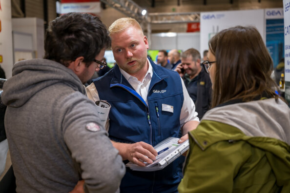 Ein Mann in einer blauen Weste erklärt zwei Personen auf einer belebten Indoor-Veranstaltung oder Messe etwas auf einem Tablet. Alle drei sind auf den Bildschirm konzentriert, und im Hintergrund sind andere Teilnehmer zu sehen.