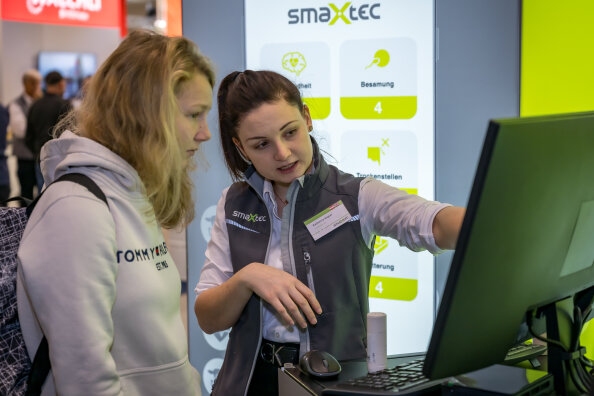Zwei Frauen stehen vor einem Computermonitor an einem Technikstand; eine Frau zeigt auf den Bildschirm und erklärt der anderen etwas. Im Hintergrund ist ein Display mit dem smaxtec-Logo und Symbolen zu sehen.