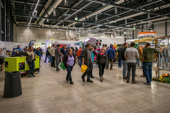 Die Menschen gehen durch eine belebte Indoor-Messe mit verschiedenen Ständen, Maschinenausstellungen und Informationsständen. Der Veranstaltungsort hat hohe Decken und eine industrielle Beleuchtung. Einige Besucher tragen Taschen, Helme oder gelbe Schutzhelme.