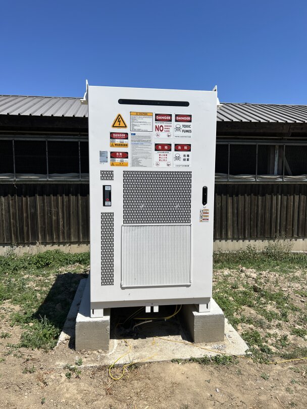 Ein großes weißes Industrie-Elektroaggregat mit Warnschildern steht auf einer Betonplattform im Freien vor einem Holzgebäude unter einem klaren blauen Himmel. Unter dem Gerät sind gelbe Kabel zu sehen.