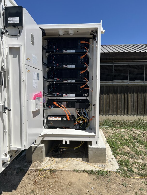 Ein Schaltschrank im Freien mit geöffneter Tür, die den Blick auf gestapelte schwarze Batteriemodule, orangefarbene und gelbe Kabel und elektrische Komponenten freigibt, die auf Betonblöcken in einer sonnigen, grasbewachsenen Fläche stehen.