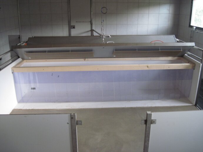 Ein großes, rechteckiges, gefliestes Becken mit einem Rahmen aus Metall und Holz, das in einem sauberen, gefliesten Raum mit weißen Wänden und einem kleinen Fenster steht, das natürliches Licht hereinlässt.