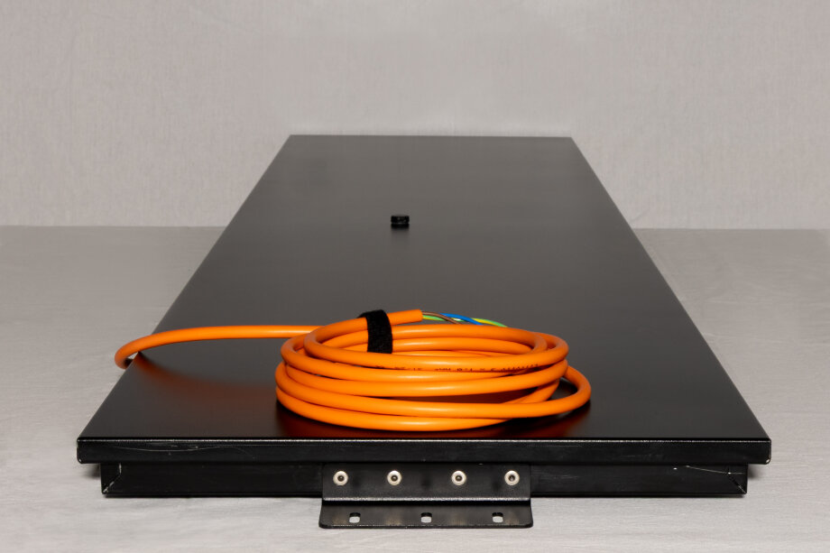 Eine schwarze, rechteckige Metallplatte liegt flach auf einem aufgerollten, orangefarbenen Kabel. Der Hintergrund ist eine schlichte hellgraue Fläche.