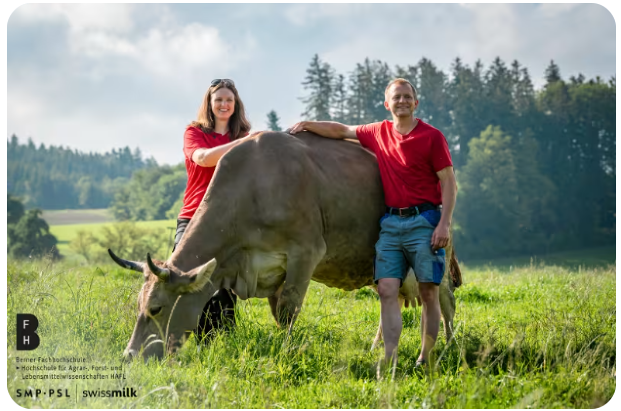 Zwei Personen in roten Hemden stehen auf einem grasbewachsenen Feld mit Bäumen im Hintergrund, lächeln und stützen ihre Hände auf eine grasende braune Kuh. Logos und Text sind in der unteren linken Ecke zu sehen.
