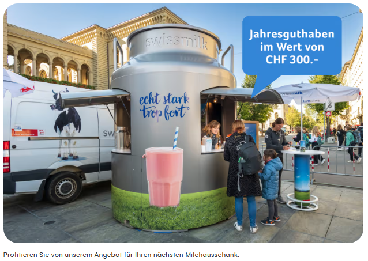 Ein riesiger Milchkannen-Kiosk mit der Aufschrift "Echt stark true fort" schenkt bei einem Strassenfest Getränke aus. Ein blaues Schild wirbt für ein Jahresguthaben von 300 Franken. Auch eine Kuh und ein Glas mit rosa Milch sind zu sehen.