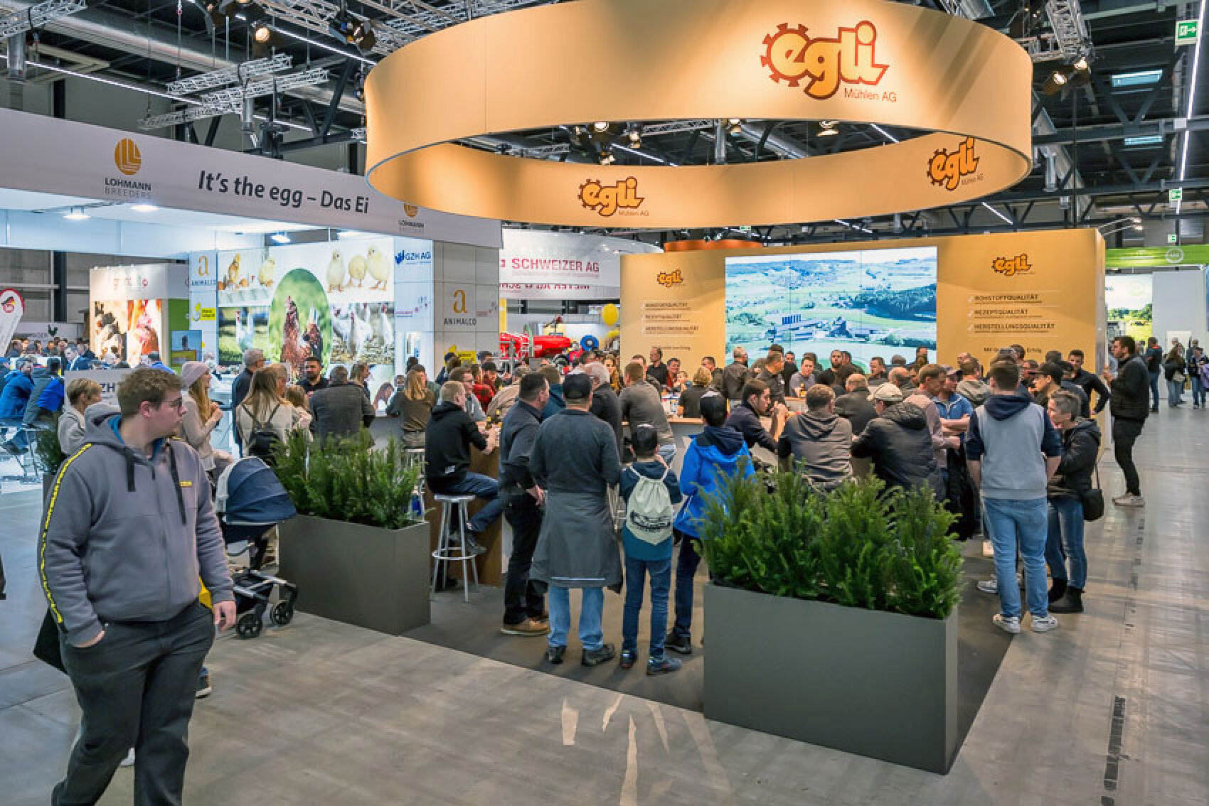 Vor einem Messestand mit gelben Bannern, Pflanzen und Ausstellungsstücken versammelt sich eine Menschenmenge. In einem großen, hell beleuchteten Kongresszentrum unterhalten sich die Besucher und betrachten die Exponate.
