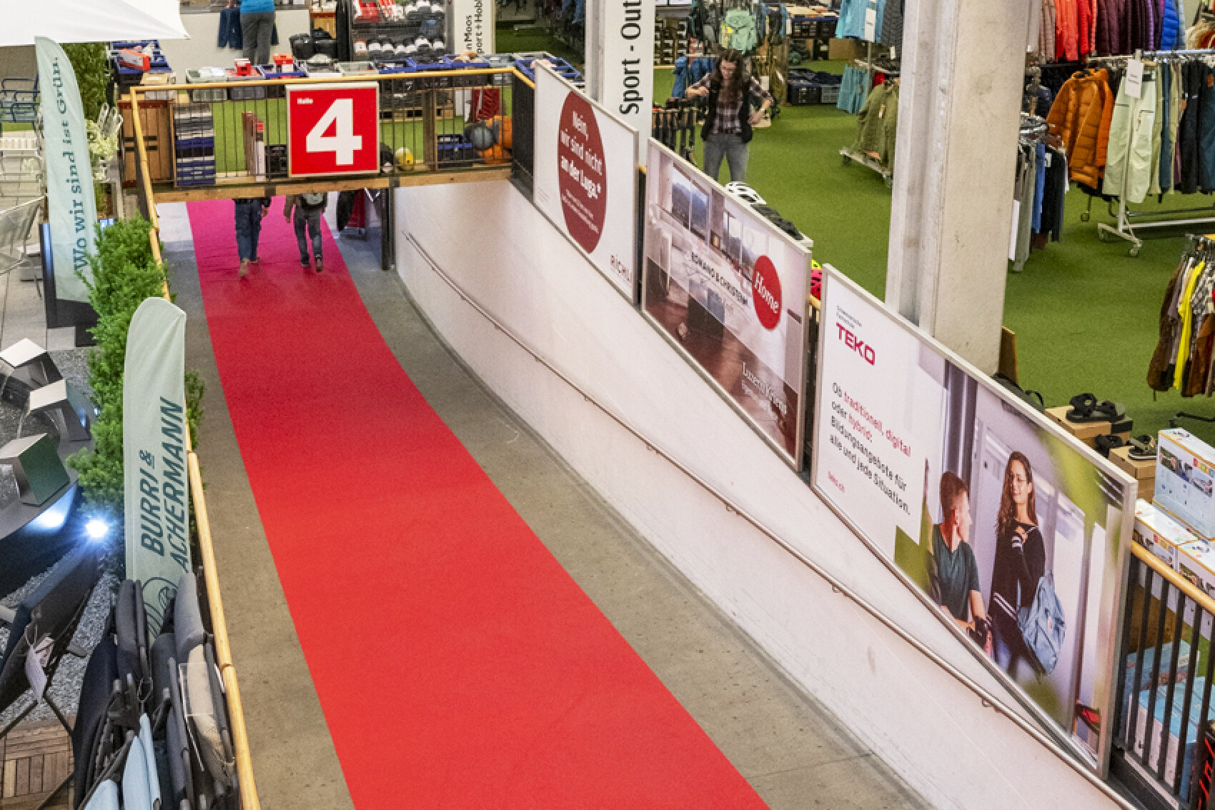 Ein roter Teppich führt durch einen Einkaufsbereich mit Kleiderständern auf grünem Boden zur Rechten und großen Plakaten an der Wand. Ein Schild mit einer weißen Zahl 4 auf rotem Grund befindet sich über dem Durchgang, in den die Leute eintreten.